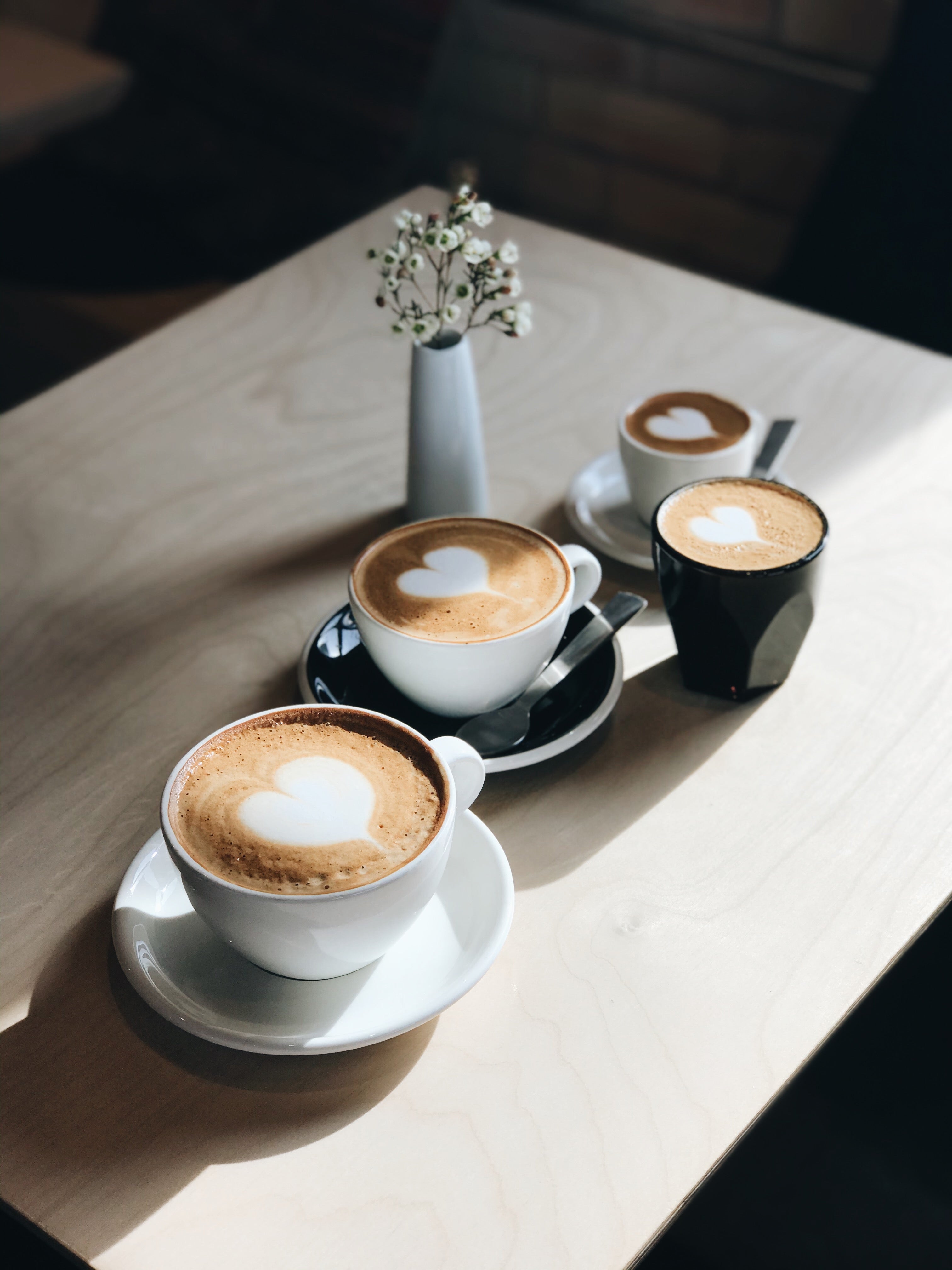 Four coffee cups with latte art on a wooden table with a vase of flowers.