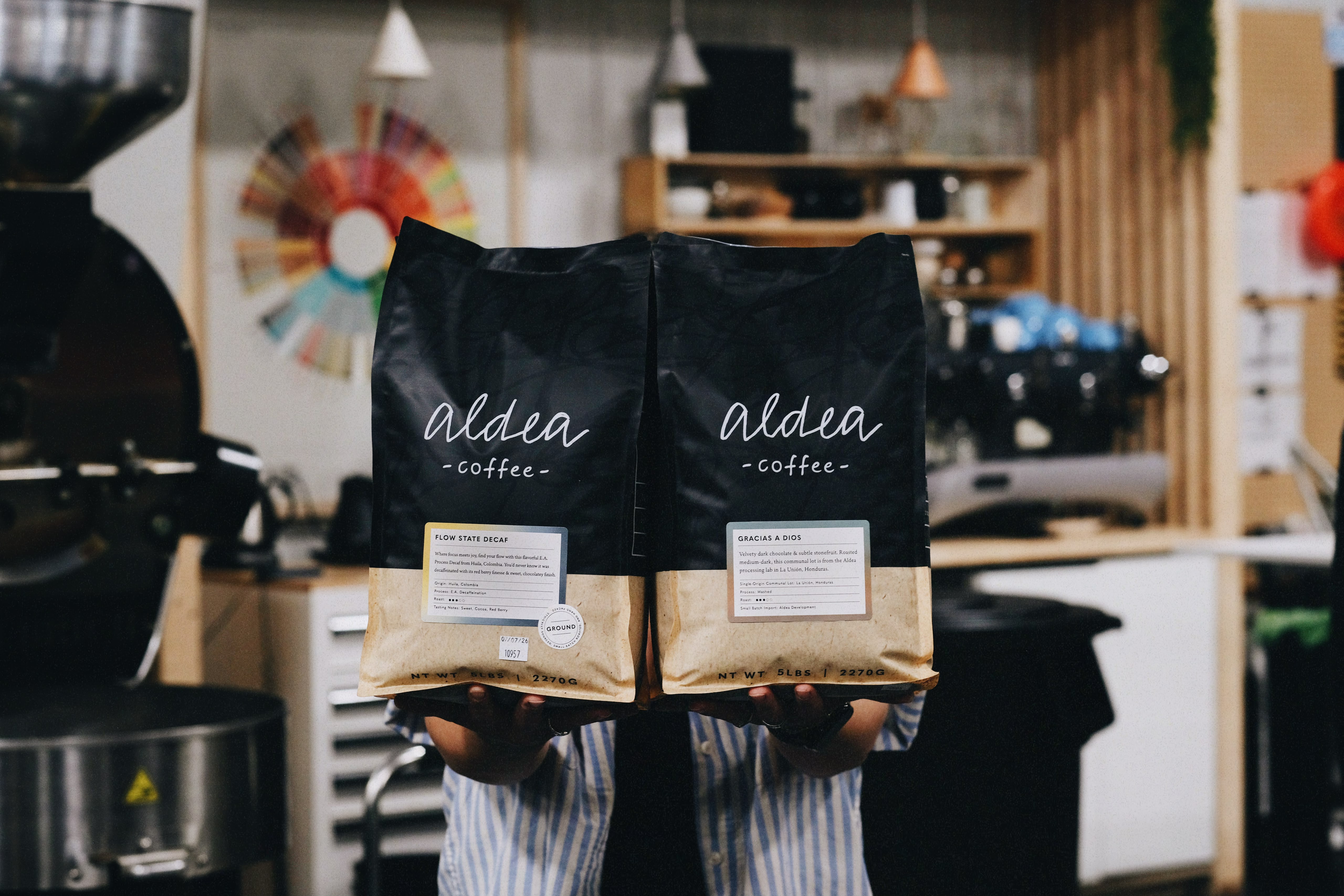 Aldea Coffee employee holds two five-pound bags of coffee, one which is Flow State Decaf and one which is Gracias A Dios, with a blurred background of a coffee roaster and coffee tasting area and flavor wheel and espresso machine.