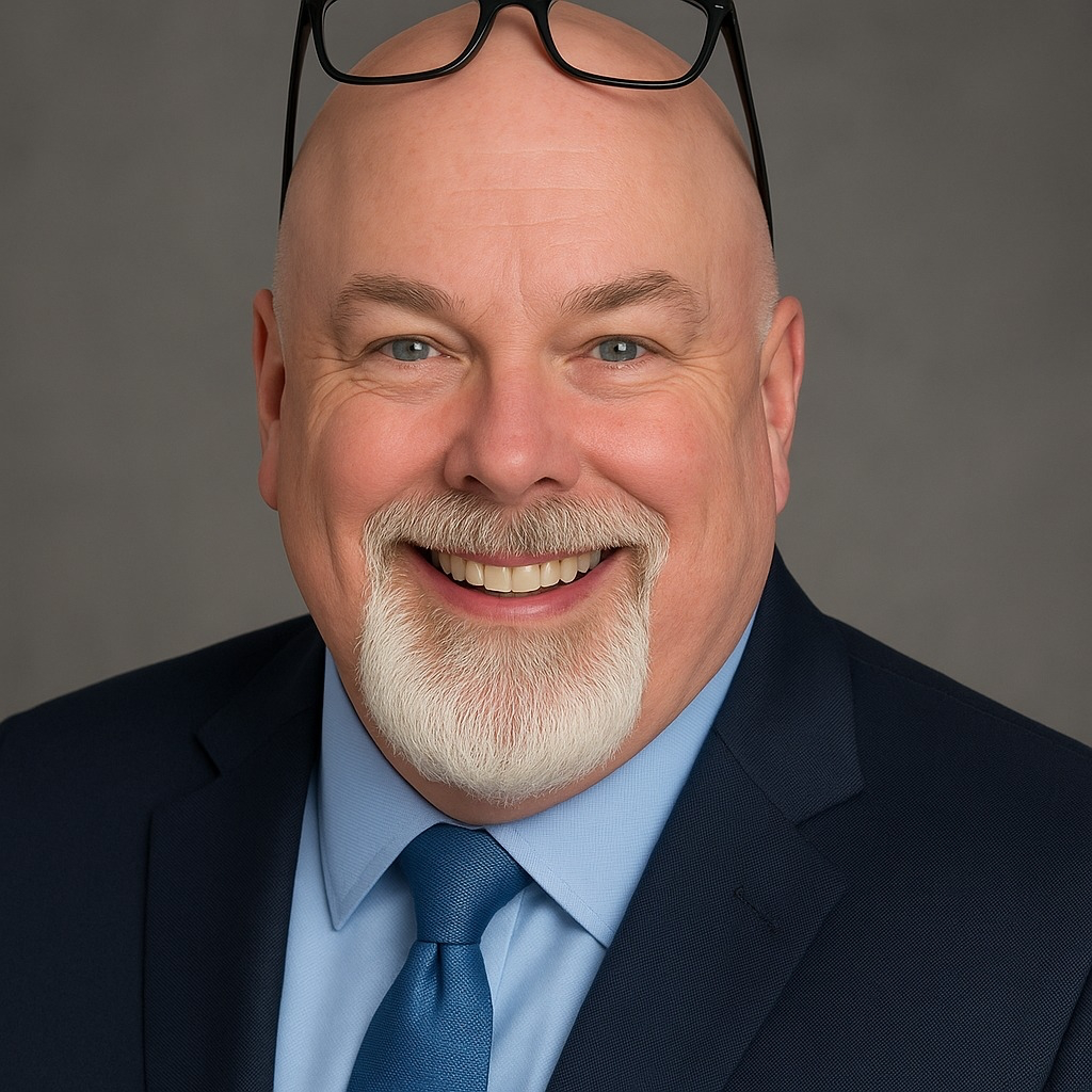 Man with a beard and glasses on his head, wearing a suit and blue tie against a gray background